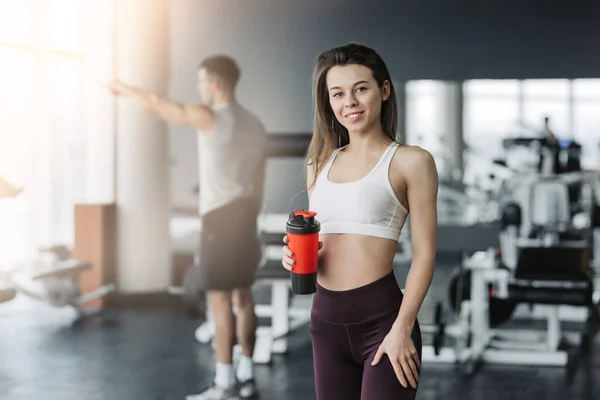 Atractiva chica deportiva sonriendo y bebiendo agua mientras está de pie en el gimnasio con el niño entrenando en segundo plano . —  Fotos de Stock