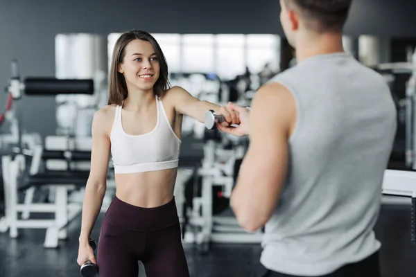 Young couple is working out at gym. Attractive woman and handsome muscular man trainer are training in light modern gym. Beautiful girl squats with dumbells under the supervision of the coach.