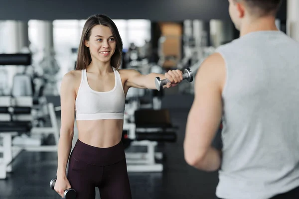 Un jeune couple s'entraîne au gymnase. Attrayant femme et beau musclé homme entraîneur s'entraînent dans la salle de gym moderne légère. Belle fille squats avec des haltères sous la supervision de l'entraîneur . — Photo