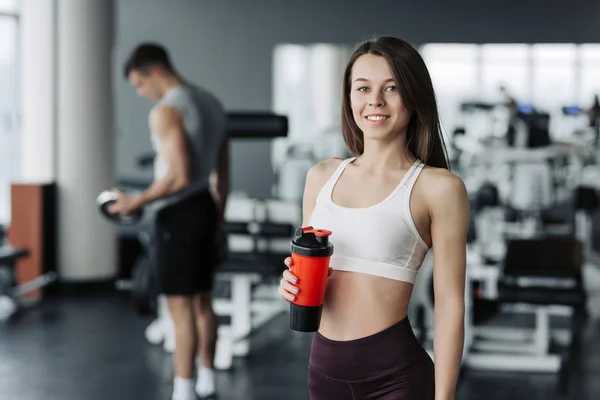 Attrayant sport fille souriant et boire de l'eau tout en se tenant à la salle de gym avec le garçon de formation sur fond . — Photo