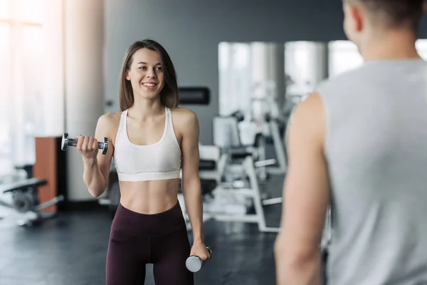 Jeune belle femme faisant des exercices avec haltère dans la salle de gym. Content que la fille souriante apprécie son processus d'entraînement. Elle travaille dur. Son formateur se penche sur ce processus . — Photo