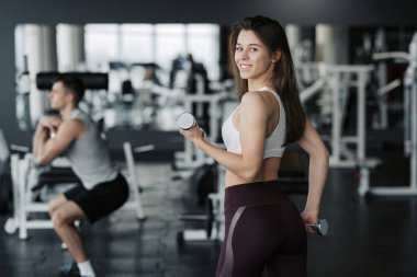 Spor salonunda ağırlık kaldıran genç bir bayan spor giyer ve arka planda erkek arkadaşı olur..