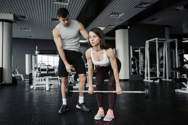 Personal trainer helping a young woman squats with dumbells in a gym.