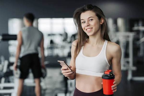 Όμορφη νεαρή αθλητική γυναίκα σε αθλητικά είδη και ακουστικά κρατά ένα μπουκάλι νερό, ακούγοντας μουσική χρησιμοποιώντας ένα τηλέφωνο και χαμογελώντας, στο γυμναστήριο. — Φωτογραφία Αρχείου