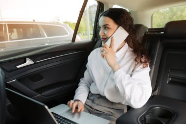 Arka koltukta dizüstü bilgisayarla oturan ve telefonla konuşurken pencerenin dışına bakan lüks bir arabayla ofise seyahat eden kadın girişimci.