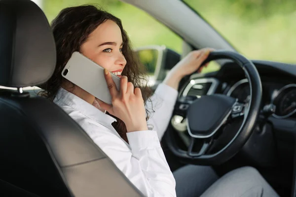 Tillbaka syn på en attraktiv ung affärskvinna tittar över axeln och pratar telefonen när du kör bil. — Stockfoto