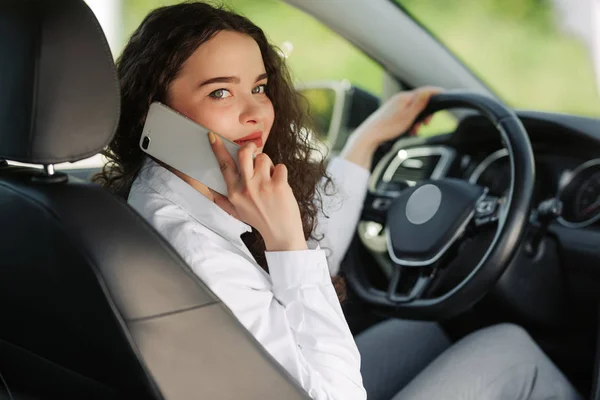 Tillbaka syn på en attraktiv ung affärskvinna tittar över axeln och pratar telefonen när du kör bil. — Stockfoto