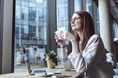 Sütlü kahve içme ve modern kentsel kafede dizüstü bilgisayar üzerinde çalışan genç kadın 