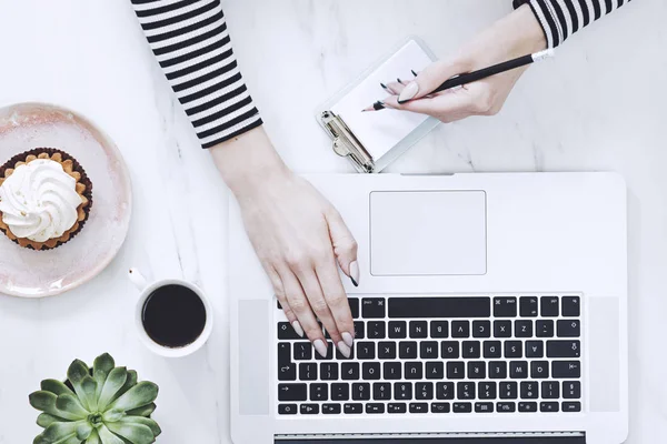 The top view of marble desk with woman hands, cup of coffee, succulent, cake and notes. Concept for social media, bussines, freelance work. Home office concept.