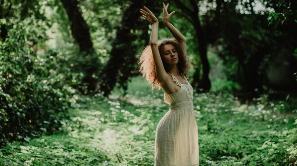 Tender curly woman in linen dress dancing in green forest