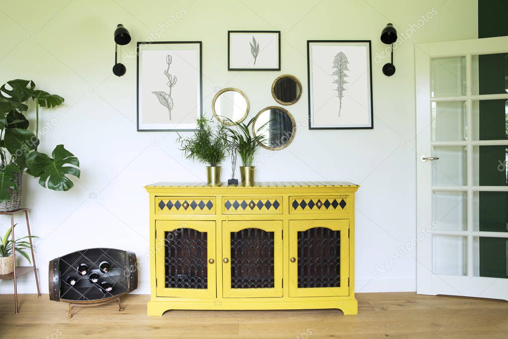 Sunny and bright living room with yellow commode, plants, mirrors and art frames