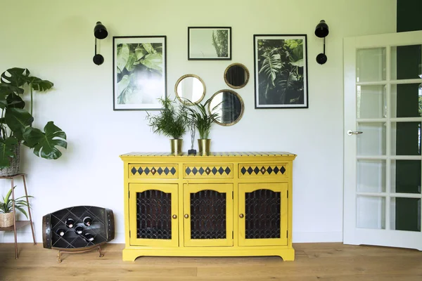 Sunny and bright living room with yellow commode, plants, mirrors and art frames