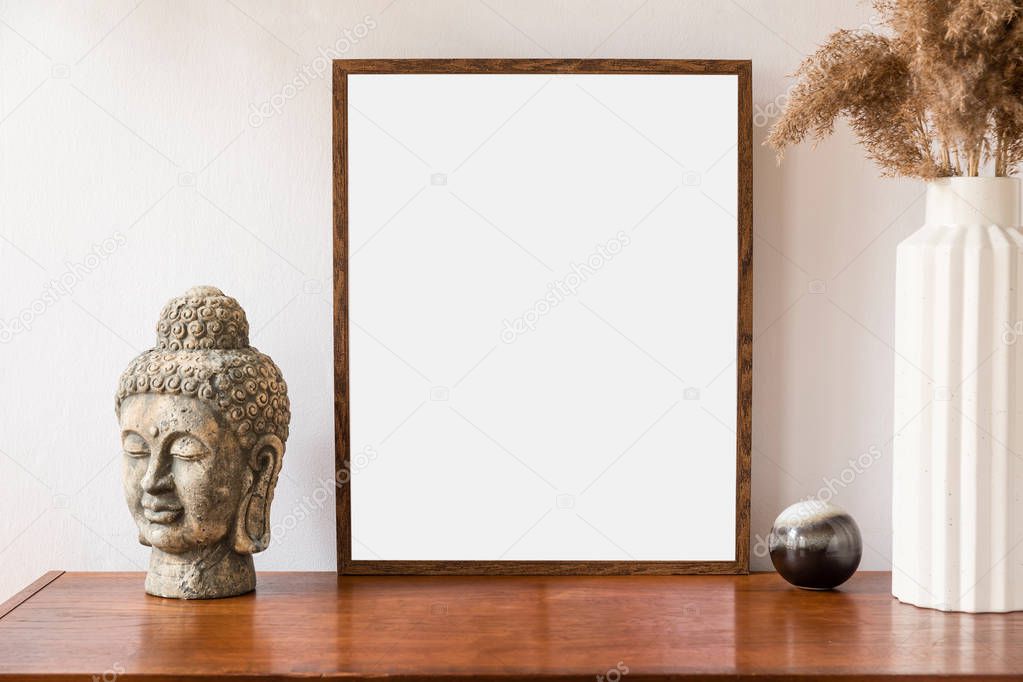 Stylish room of home interior with brown mock up frame with vintage accessories, buddha head and flowers in vase. Cozy home decor. Minimalistic concept. Retro composition of living space.