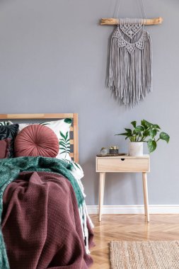 Tasarım ahşap gece masası, gri makrame, zarif aksesuarlar ve bitki ile yatak odası Lüks iç. Güzel çiçekli yatak çarşafları, battaniye ve yastıklar ile oturma odası Modern ev dekorasyonu. Şablon