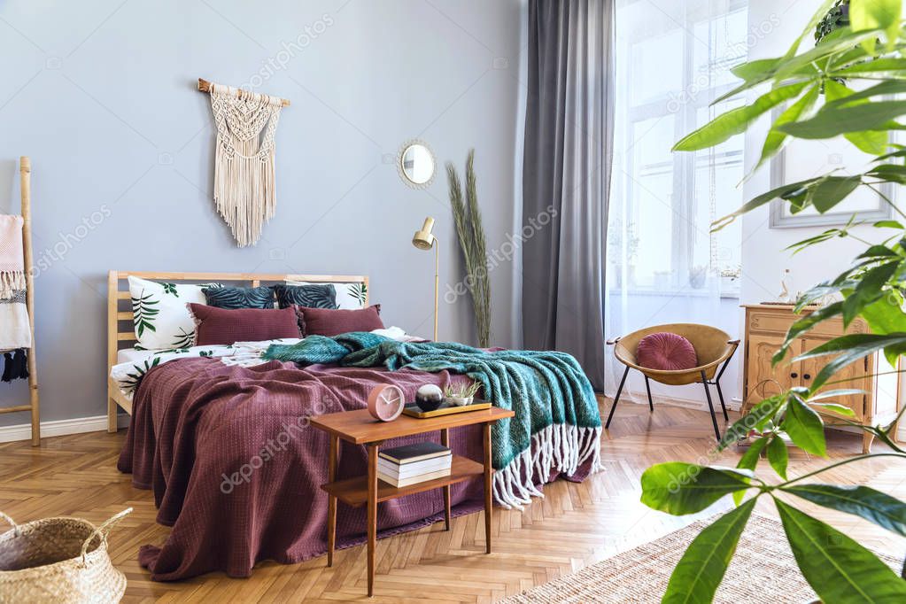 Stylish and luxury interior of bedroom with design furnitures, honey yellow armchair, gray macrame on the walls and elegant accessories. Beautiful bed sheets, blankets and pillows. Warm home decor.