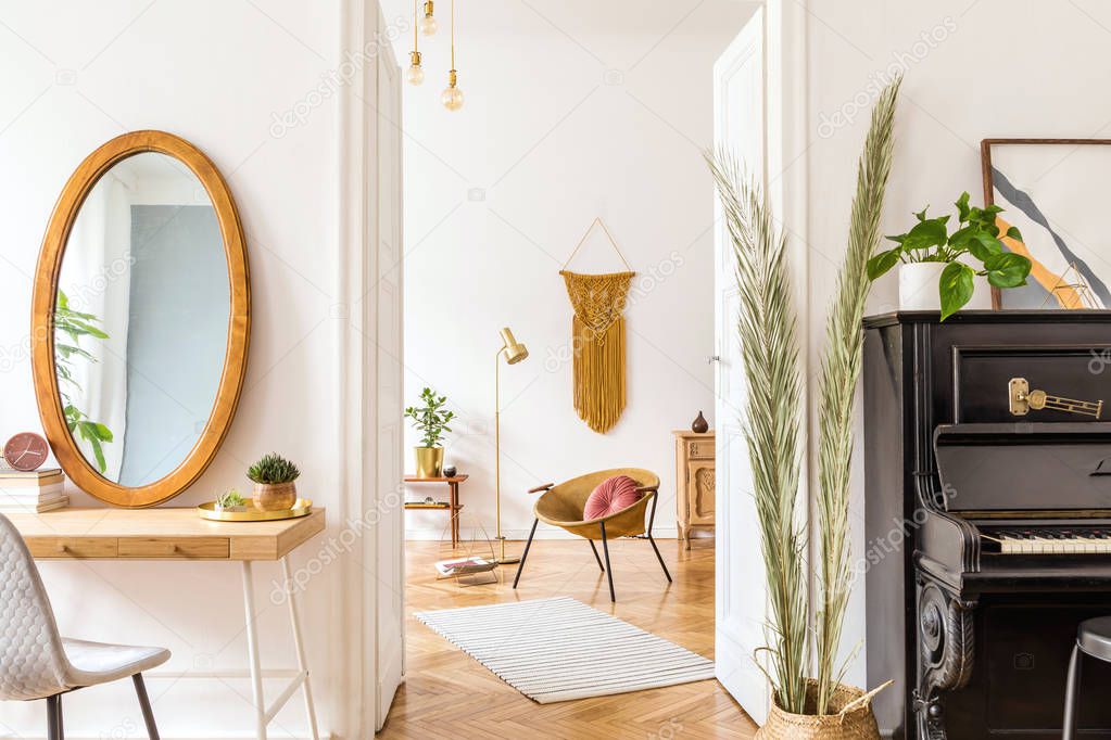 Stylish interior of apartment with gold armchair, wooden mirror, frames, design furnitures, piano, accessories, plants, gold lamp and macrame on the white wall. Modern home decor of living room. 