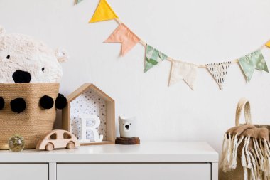 Beyaz raf, oyuncak ayı, ahşap oyuncaklar ve ev ile şık İskandinav kreş iç. Beyaz arka plan duvarına pamuklu bayraklar ve doğal sepet asmak. Rahat ve güneşli çocuk odası.