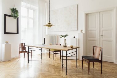 Tasarım paylaşım masası, sandalye, altın pedant lamba, soyut resimler ve şık aksesuarlar ile güneşli ve zarif yemek odası iç. Vazodaki tropikal yapraklar. Eklektik dekor. Kahverengi ahşap parke.