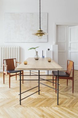 Eklektik ve zarif yemek odası iç tasarım paylaşım masası, sandalyeler, altın pedant lamba, soyut resimler ve şık aksesuarlar. Vazodaki tropikal yapraklar. Eklektik dekor. Kahverengi ahşap parke.