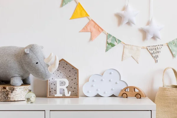 The modern scandinavian newborn baby room with plush rhino, design boxes, wooden toys, cloud and natural basket. Hanging cotton flags and stars on the white background wall. Stylish and cozy interior.