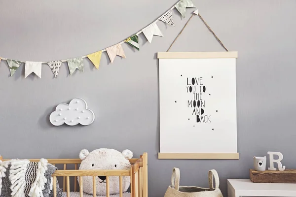 Stylish and cute scandinavian decor of newborn baby room with mock up poster, natural toys, hanging decor flags and cloud, wooden cradle, basket for accessories and teddy bears. Grey walls .