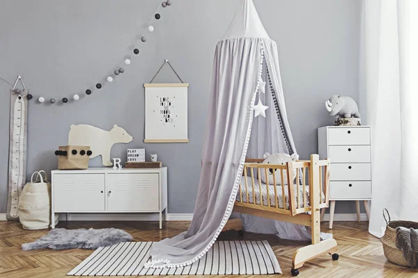 Stylish and cute scandinavian decor of newborn baby room with mock up poster , white design furnitures, natural toys, hanging grey canopy with wooden cradle, pillows, accessories and teddy bears.