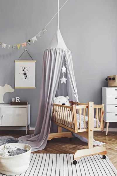 Stylish and cute scandinavian decor of newborn baby room with mock up poster , white design furnitures, natural toys, hanging grey canopy with wooden cradle, pillows, accessories and teddy bears.