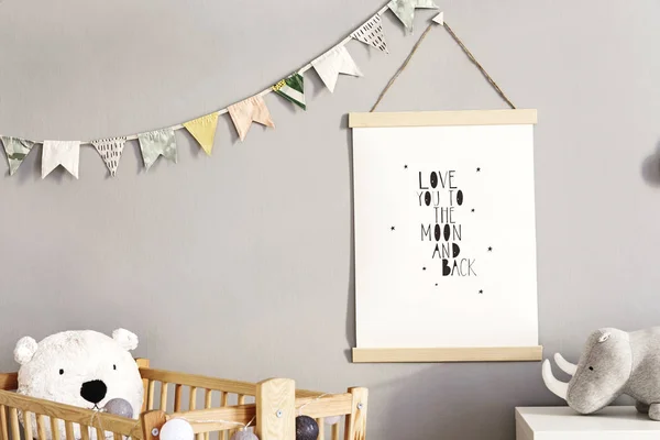 Stylish and cute scandinavian decor of newborn baby room with mock up poster, natural toys, hanging decor flags and cloud, wooden cradle, plush rhino and teddy bears. Grey walls.