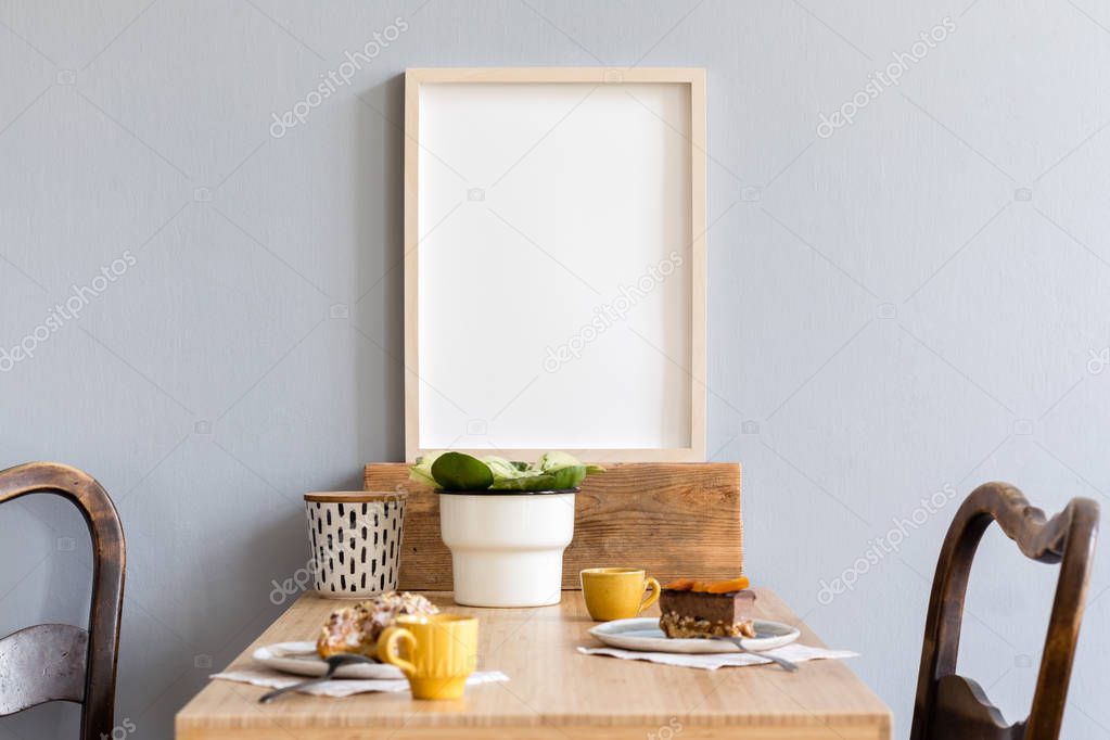 Stylish and retro interior of kitchen space with small wooden table with mock up photo frame, plant, box, design cups, furnitures and tasty dessert. Scandinavian room decor with kitchen accessories.
