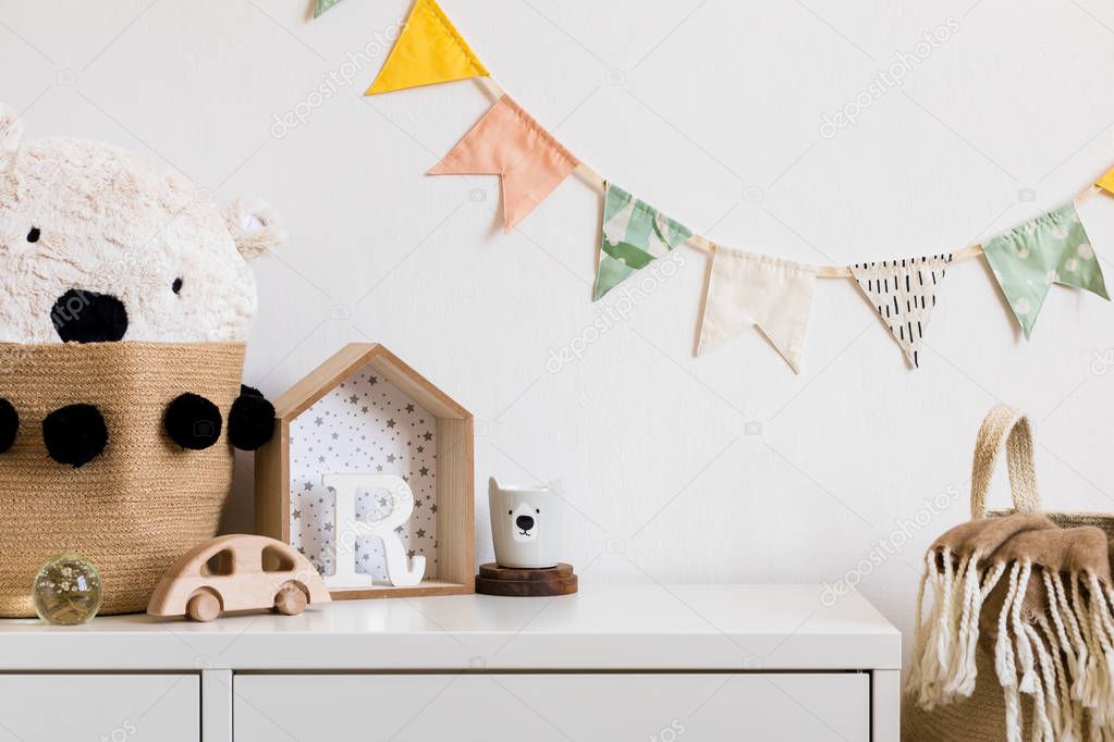 Stylish scandinavian nursery interior with white shelf, teddy bear, wooden toys and house. Hanging cotton flags and natural basket on the white background wall. Cozy and sunny childroom.