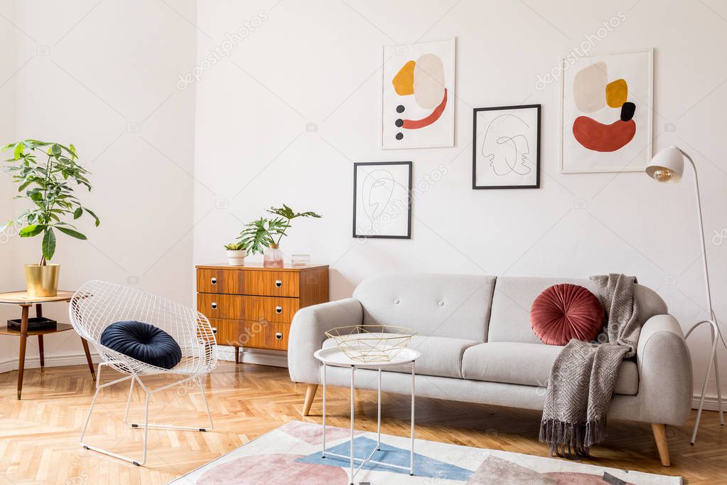 Stylish vintage decor in a spacious flat interior with design grey sofa, armchair, retro commode and posters on the wall. Brwon wooden parquet, stylish carpet and plants. Bright living room.