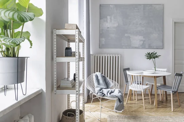 Modern nordic living room with design furniture, family table, plant, white bookstand on the wall. Brown wooden parquet. Abstract painting on the white wall. Concept of minimalistic interior.