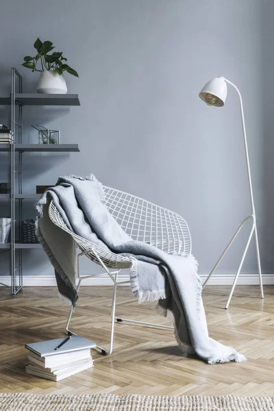 Stylish nordic living room with design armchair with blanket, white lamp and bookstand on the grey wall. Brown wooden parquet. Concept of minimalistic space.