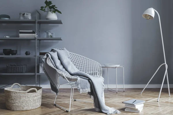 Stylish nordic living room with design armchair with blanket, white lamp and bookstand on the grey wall. Brown wooden parquet. Concept of minimalistic space.