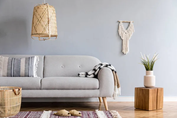 Stylish and design home interior of living room with gray sofa, wooden cube, pillow, rattan lamp and basket, macrame, carpet and elegant accessories. Stylish home decor. Template. Gray wall