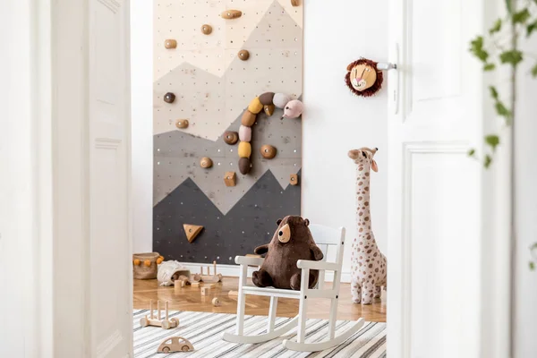 Stylish interior design of childroom with modern climbing wall for kids, design furnitures, soft toys, teddy bear on armchair and cute children\'s accessories. Scandinavian playroom decor. Template.