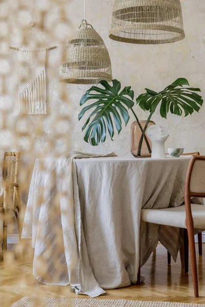 stock image Stylish dining room interior with wooden table, design chairs, rattan pendant lamps, tropical leafs in vase and elegant decoration. Wabi sabi concept. Template.