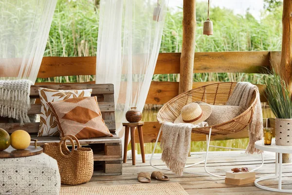 Interior design of summer gazebo by the lake with stylish rattan armchair, coffee table, sofa, pillows, plaid and elegant accessories in modern decor. Summer vibes. Chillout. Template.