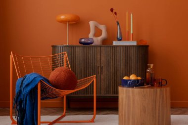 Eklektik ve modern oturma odasında heykel gibi bir hava var. Turuncu tel sandalye, yuvarlak ahşap masa ve turuncu bir duvara karşı duran kalın aksesuarlar..