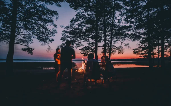 Grupo de jóvenes amigos felices sentados junto al fuego en la playa de verano, asando salchichas y bebiendo cerveza, hablando y divirtiéndose — Foto de Stock