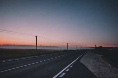 Sabah geldi. Turuncu güneşin doğacağını puslu bir alan, yol asfalt ve orman, karanlık ağaçlar.