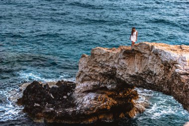 Woman in white dress on the beautiful natural rock arch near of Ayia Napa, Cavo Greco and Protaras on Cyprus island, Mediterranean Sea. Legendary bridge lovers. clipart