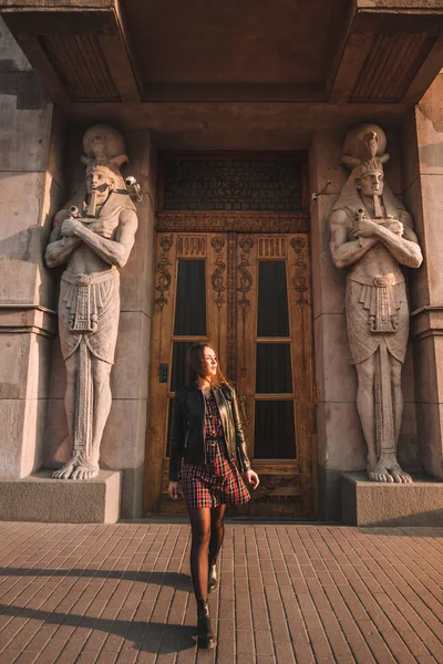 Attractive girl with blond hair in a dress in a cage and a black leather jacket in the sunlight goes forward to the camera. behind the "Egyptian house" with the statues of the pharaohs at the entrance — Stock Photo, Image