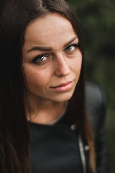 Retrato de uma menina bonita com cabelos longos, olhos grisalhos em uma jaqueta de couro preto olhando diretamente para a câmera. Floresta em segundo plano . — Fotografia de Stock