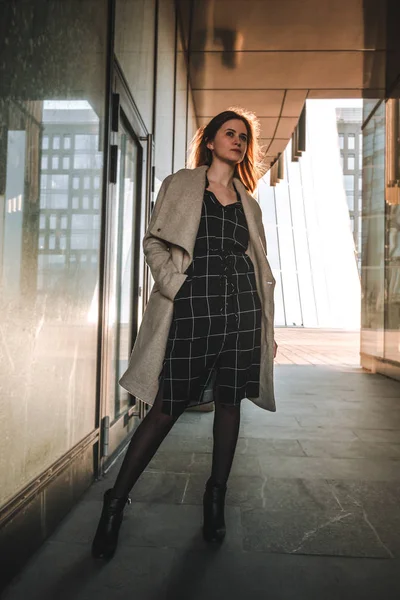 Photo of a girl in a coat and dress against the background of a modern business center, backlight — Stock Photo, Image