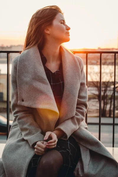 Kavkazská hnědá dívka v béžovém kabátě sedí ve větru a dívá se stranou. světlo slunce za — Stock fotografie
