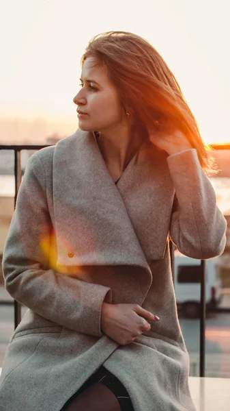 Chica de pelo castaño caucásico en un abrigo beige endereza el cabello en el viento. atardecer detrás —  Fotos de Stock