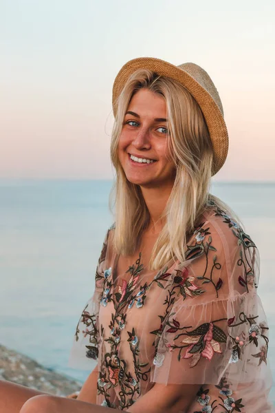 Mladá hezká žena s blond vlasy v růžové košili a klobouk sedí na kamenech v blízkosti moře při východu slunce — Stock fotografie