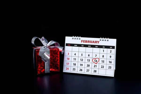 Calendar with red written heart highlight on February 14 of Saint Valentines day as reminder, not forget for couples to express their love with gift box on black background. Concept of Valentine's day