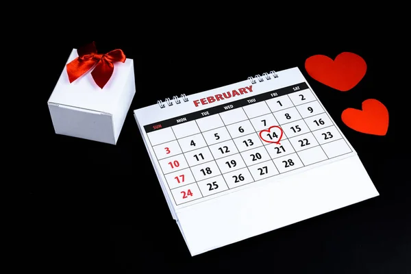 Calendar with red written heart highlight on February 14 of Saint Valentines day as reminder for couples to express love with heart shape, gift box on black background. Concept of Valentine\'s day